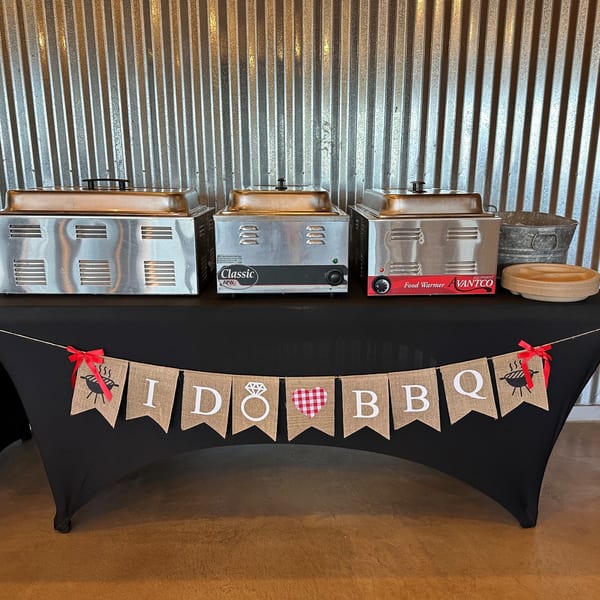Three food warmers on a black-draped table with an "I DO BBQ" banner, against a corrugated metal wall.