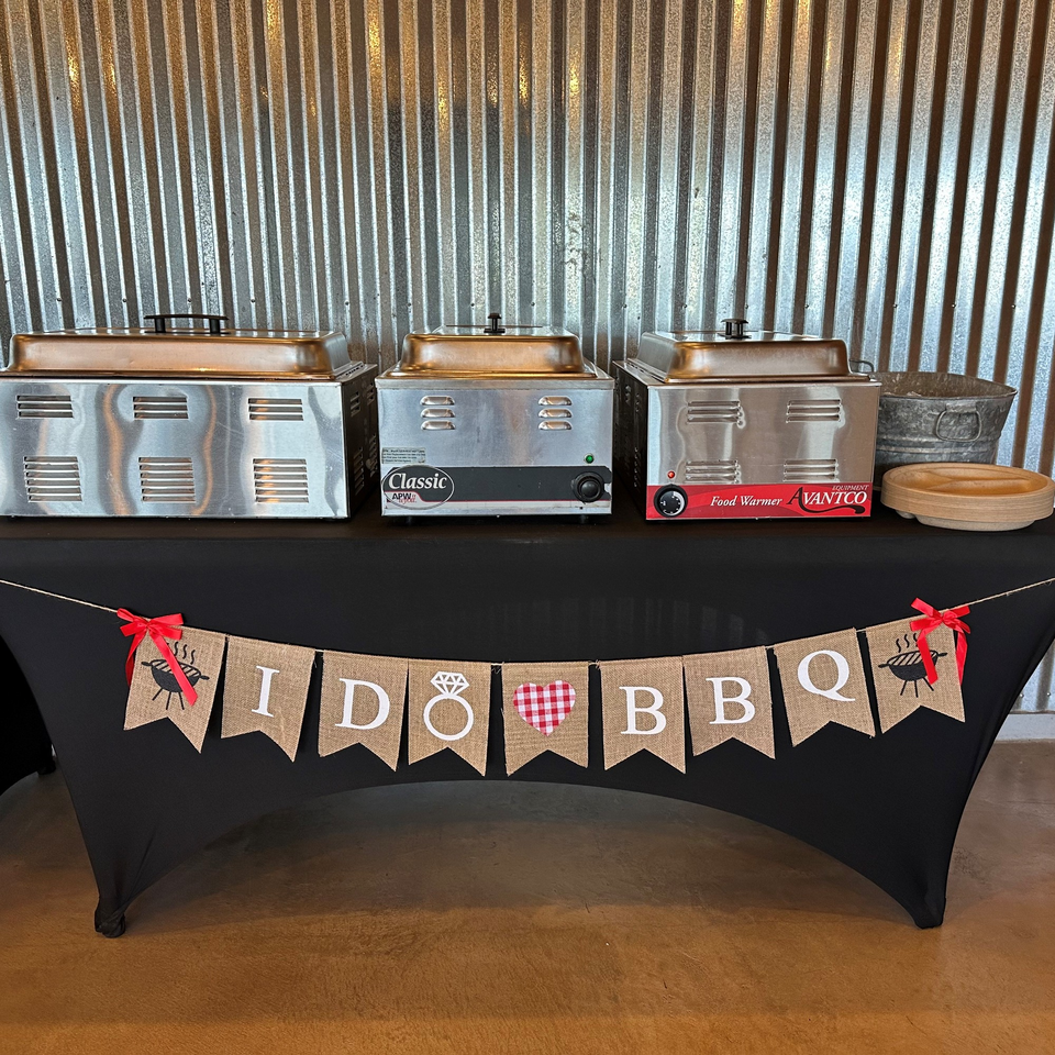Three food warmers on a black-draped table with an "I DO BBQ" banner, against a corrugated metal wall.