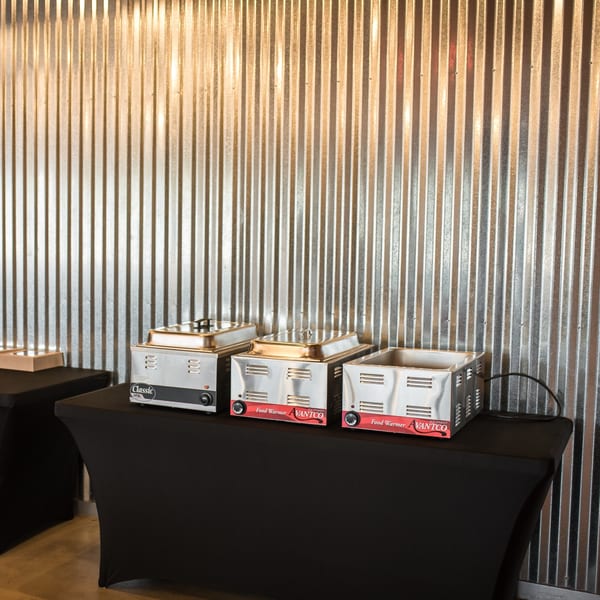 Three stainless steel food warmers on black-draped tables against a corrugated metal wall.
