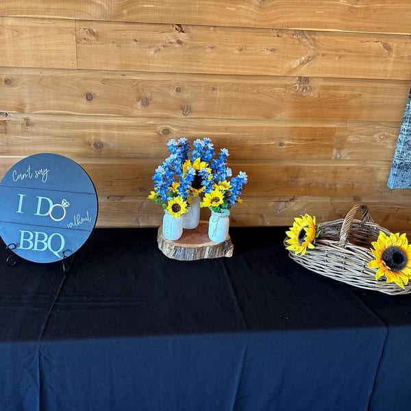 A blue "Can't say I Do without BBQ" sign, mason jars with blue and yellow flowers, and a wicker basket.