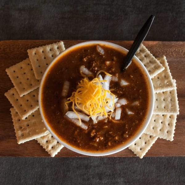 Meyer's Elgin Smokehouse chili with shredded cheese, diced onions, and crackers on a wooden board.